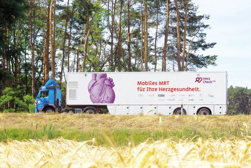 Ein blauer Lkw steht auf einem Feld, umgeben von Bäumen. An der Seite des Fahrzeugs ist ein Bild eines Herzens sowie der Text "Mobiles MRT für Ihre Herzgesundheit" zu sehen.