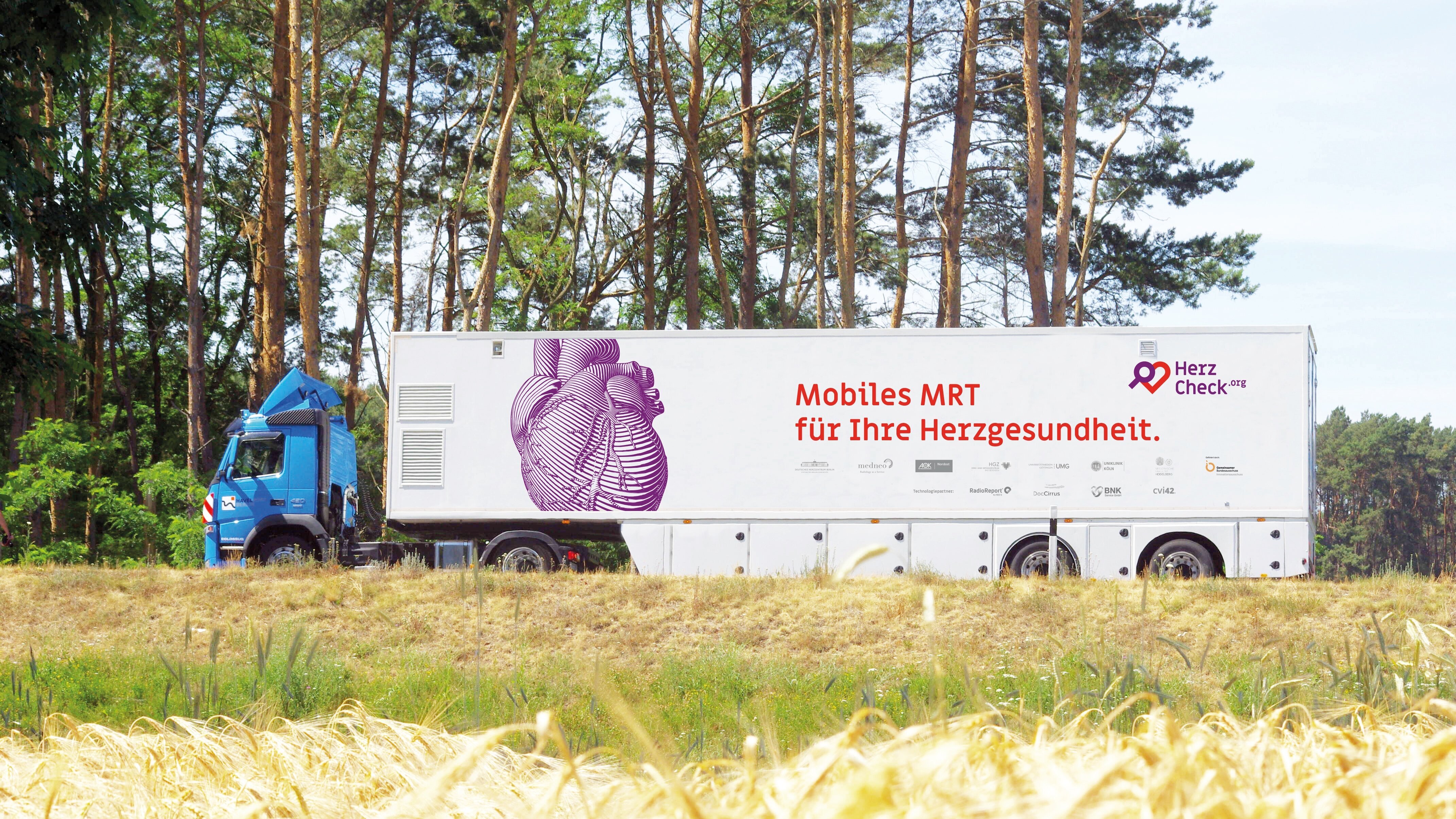 Ein blauer Lkw steht auf einem Feld, umgeben von Bäumen. An der Seite des Fahrzeugs ist ein Bild eines Herzens sowie der Text "Mobiles MRT für Ihre Herzgesundheit" zu sehen.