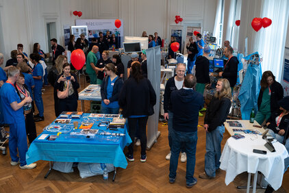 Eine belebte Veranstaltungsfläche mit Menschen in medizinischer Kleidung. Einige Personen unterhalten sich, während andere Informationsmaterial an Tischen anschauen. Rote Luftballons schweben im Raum. Der Fokus liegt auf dem Austausch von Informationen und der Präsentation medizinischer Geräte.