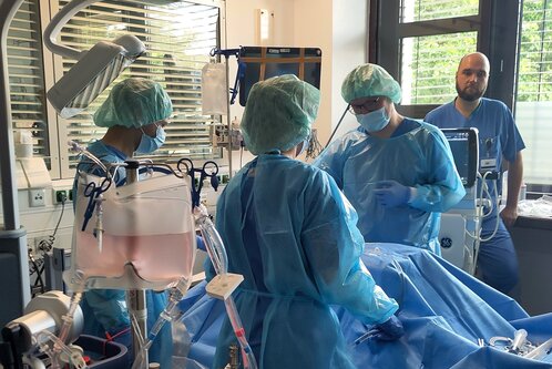 Im Bild befinden sich mehrere medizinische Fachkräfte in Schutzkleidung, die an einem Patienten arbeiten. Sie nutzen verschiedene medizinische Geräte, um eine Behandlung in einem Krankenhauszimmer durchzuführen. Ein weiterer Mitarbeiter beobachtet die Situation im Hintergrund.