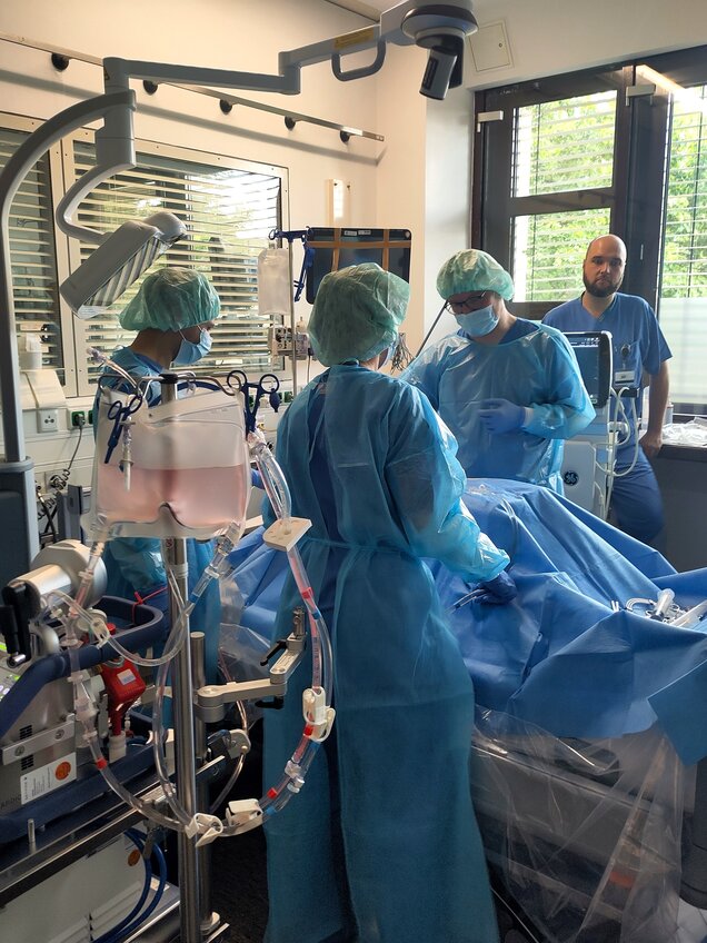 Im Bild befinden sich mehrere medizinische Fachkräfte in Schutzkleidung, die an einem Patienten arbeiten. Sie nutzen verschiedene medizinische Geräte, um eine Behandlung in einem Krankenhauszimmer durchzuführen. Ein weiterer Mitarbeiter beobachtet die Situation im Hintergrund.