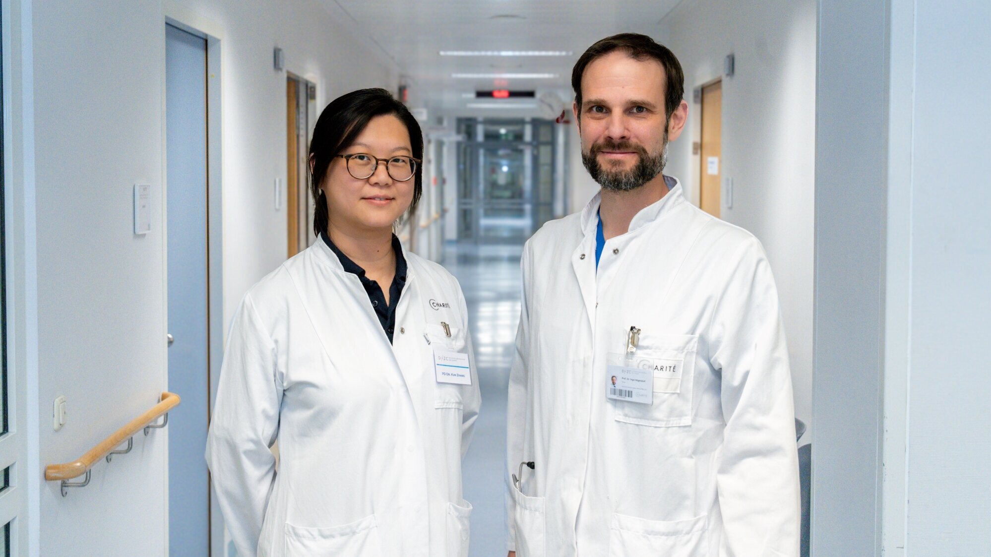 Zwei Ärzte in weißen Kitteln stehen in einem Krankenhausflur. Die Person links trägt eine Brille und lächelt, während die Person rechts eine leicht neutrale Miene hat. Im Hintergrund sind Krankenhauszimmer und Lichter sichtbar.