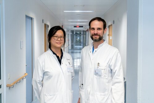 Zwei Ärzte in weißen Kitteln stehen in einem Krankenhausflur. Die Person links trägt eine Brille und lächelt, während die Person rechts eine leicht neutrale Miene hat. Im Hintergrund sind Krankenhauszimmer und Lichter sichtbar.