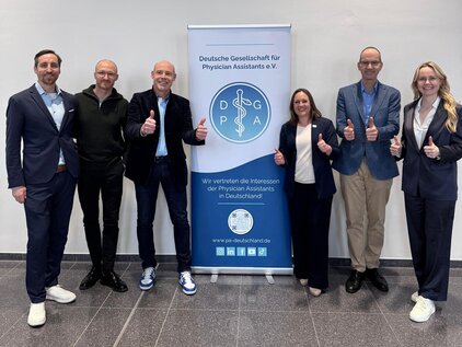 Sechs Personen in formeller Kleidung stehen vor einem Banner der Deutschen Gesellschaft für Physician Assistants. Alle zeigen Daumen nach oben und lächeln. Das Banner betont die Vertretung der Interessen von Physician Assistants in Deutschland.
