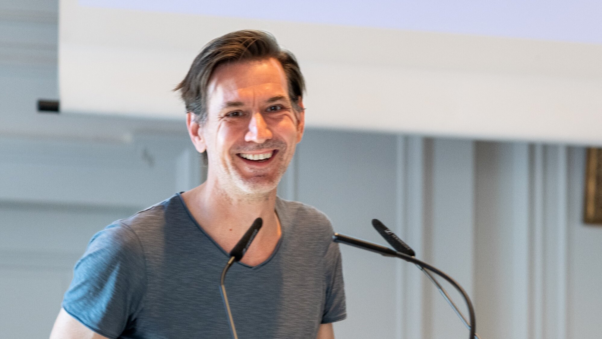 Ein lächelnder Mann steht an einem Podium mit dem Logo des Deutschen Herz-Zentrums der Charité. Er trägt ein schlichtes, graues T-Shirt und ist anscheinend in eine Präsentation oder Rede vertieft. Im Hintergrund sind unscharfe Elemente eines Konferenzraums sichtbar.