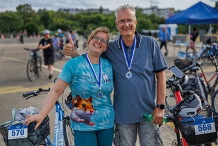 Ein Mann und eine Frau stehen lächelnd nebeneinander auf einem Fahrradparkplatz. Beide tragen eine Medaille und zeigen den Daumen hoch. Die Frau hat ein T-Shirt mit einem Hamstermotiv. Im Hintergrund sind weitere Radfahrer und ein blauer Pavillon sichtbar.