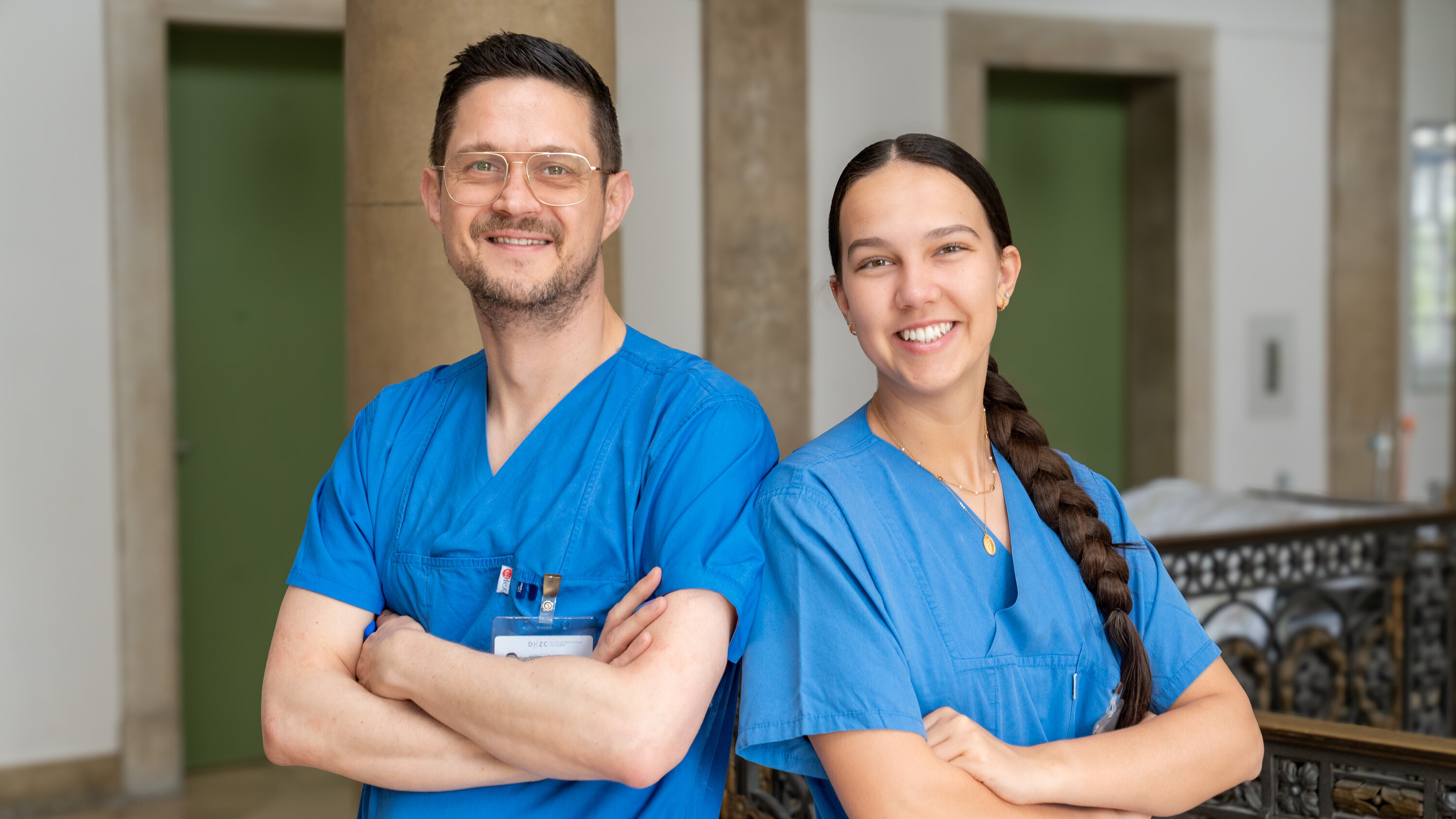 Zwei medizinische Fachkräfte in blauen Kitteln stehen lächelnd mit verschränkten Armen in einer Klinik. Der Mann hat Brille und kurze Haare, die Frau hat lange, geflochtene Haare. Der Hintergrund zeigt eine helle, moderne Krankenhausumgebung.