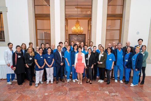 Eine Gruppe von etwa 30 Personen steht im Freien vor einem Gebäude mit großer Eingangstür und Kronleuchter. Die Personen tragen verschiedene Arbeitskleidungen, hauptsächlich medizinische Uniformen in Blau und Weiß. Im Vordergrund steht eine Frau in einem pinken Kleid.