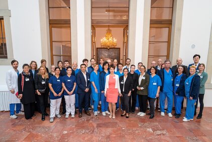 Eine Gruppe von etwa 30 Personen steht im Freien vor einem Gebäude mit großer Eingangstür und Kronleuchter. Die Personen tragen verschiedene Arbeitskleidungen, hauptsächlich medizinische Uniformen in Blau und Weiß. Im Vordergrund steht eine Frau in einem pinken Kleid.