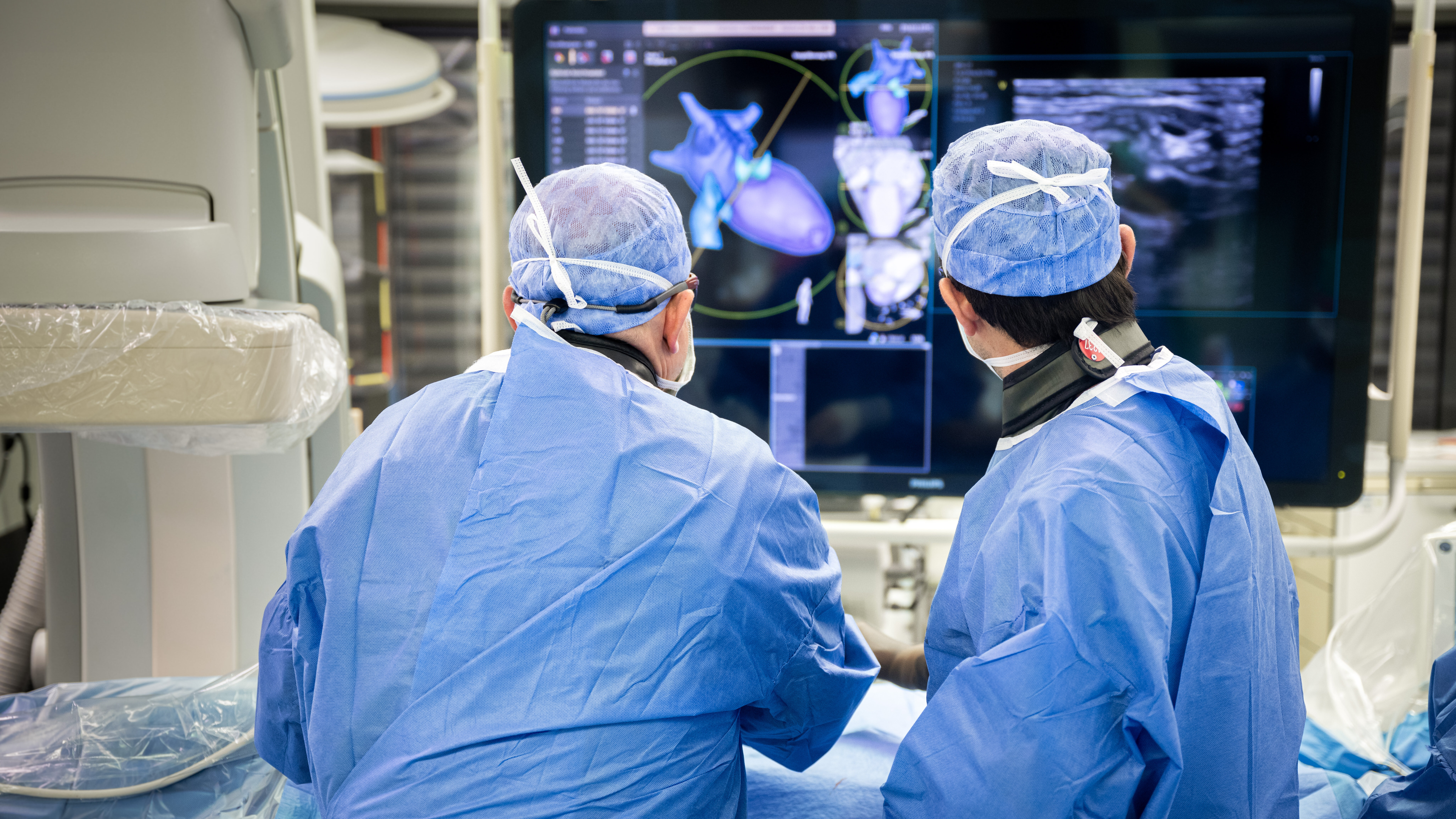 Zwei Ärzte in blauer OP-Kleidung betrachten konzentriert einen Monitor, der medizinische Bildgebungen zeigt. Die Bilder scheinen Herzstrukturen und andere anatomische Details darzustellen. Der Raum wirkt hochmodern und ist gut ausgestattet für medizinische Eingriffe.