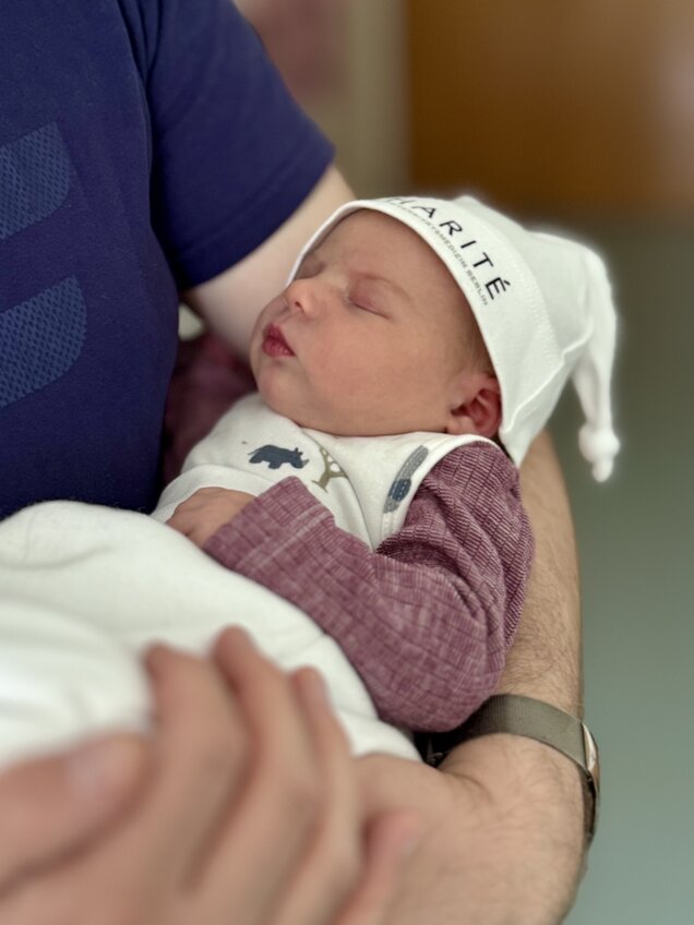 Ein Neugeborenes schläft friedlich in den Armen einer Person. Das Baby trägt eine weiße Mütze mit der Aufschrift "CARITÉ" und einen Strampler. Die Person hält das Baby sanft, der Hintergrund ist unscharf und hell.