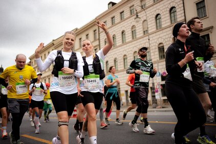 Zwei fröhliche Läuferinnen in Sportkleidung lächeln und posieren während eines Marathons. Sie tragen Wettkampfnummern und haben Wasserflaschen in den Händen. Im Hintergrund sind weitere Teilnehmer zu sehen, die den Wettkampf ebenfalls bestreiten.