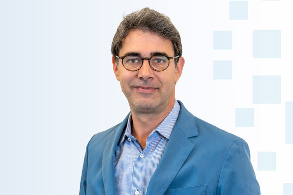 Ein Mann mit bräunlichen Haaren trägt eine Brille und einen blauen Anzug. Er hat ein freundliches Lächeln und steht vor einem hellen, geometrischen Hintergrund.