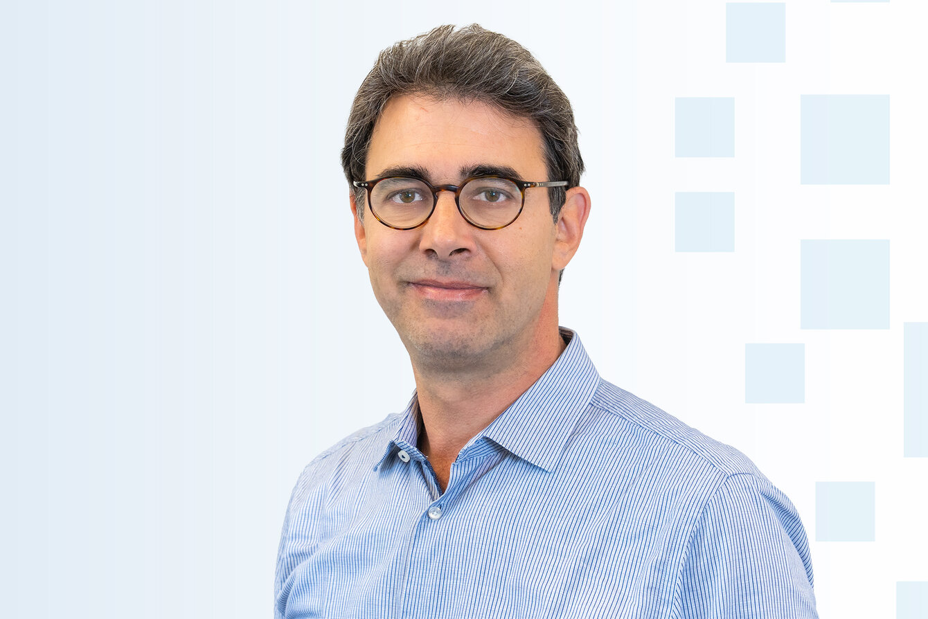 Ein Mann mit bräunlichen Haaren trägt eine Brille und einen blauen Anzug. Er hat ein freundliches Lächeln und steht vor einem hellen, geometrischen Hintergrund.