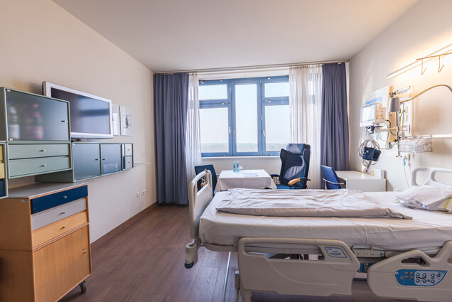 Ein Krankenhauszimmer mit einem Bett, einem Fernseher an der Wand und einem Tisch daneben. Der Raum hat große Fenster mit Vorhängen und ist mit einem Rollstuhl sowie zwei Stühlen ausgestattet. Die Wände sind hell, der Boden aus Holz.