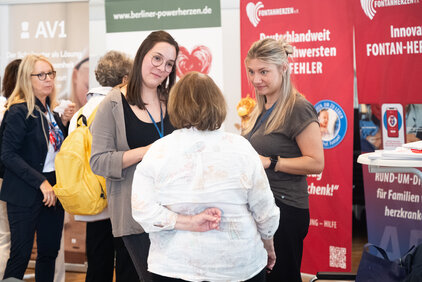 Drei Frauen stehen in einem Gespräch auf einer Veranstaltung. Eine Frau mit langen, blonden Haaren und eine mit dunklen, schulterlangen Haaren sprechen mit einer dritten Frau, die ihnen zugewandt ist. Im Hintergrund sind Informationsstände zu sehen.
