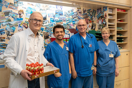 Eine Gruppe von vier Personen steht in einem Raum mit einer Pinnwand im Hintergrund, die viele Fotos zeigt. Der erste Mann trägt einen weißen Arztkittel und hält ein Tablett mit Schokolade. Die anderen drei Personen tragen blaue Arbeitskleidung und lächeln freundlich.