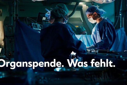 Zwei Chirurgen in einem Operationssaal führen einen Eingriff durch. Im Vordergrund steht der Text „Organspende. Was fehlt.“ Der Raum ist schwach beleuchtet und zeigt medizinische Instrumente, um die Dringlichkeit und Bedeutung von Organspenden zu betonen.