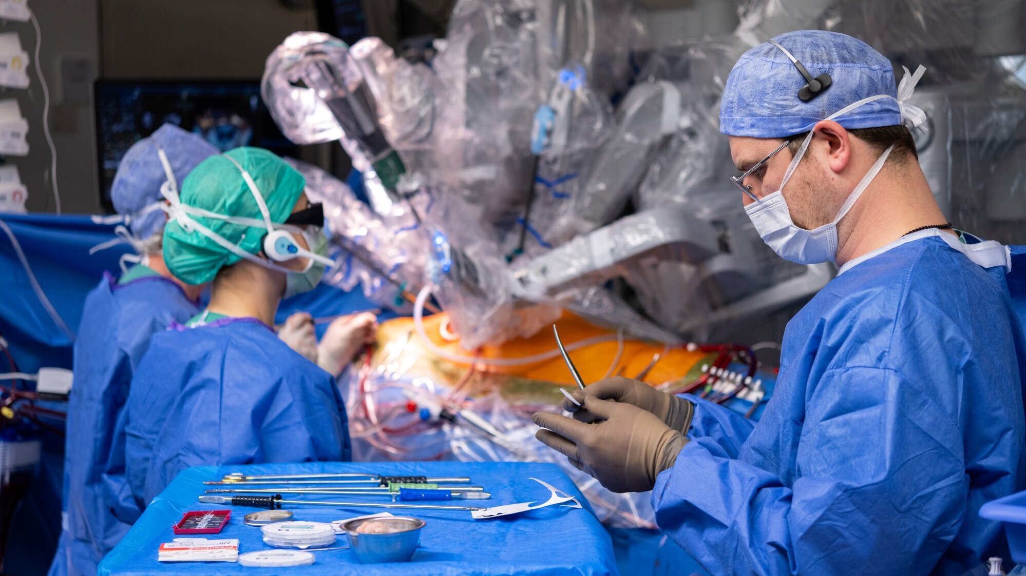 Ein Team von Chirurgen in blauen OP-Kitteln arbeitet an einem Patienten, während sie ein robotergestütztes chirurgisches System verwenden. Auf einem Tisch vor ihnen liegen verschiedene chirurgische Instrumente und Materialien. Die Atmosphäre ist konzentriert und professionell.