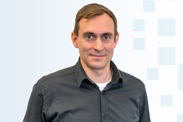 Ein Mann mit kurzen, braunen Haaren und einem neutralen Gesichtsausdruck trägt ein dunkelblaues Hemd. Er steht vor einem hellen, grafischen Hintergrund. Der Fokus liegt auf seiner Person.