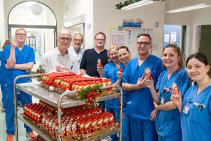 Eine Gruppe von sieben Personen in blauen Kitteln lächelt vor einem Wagen voller Nikolausfiguren. Sie halten einige der Figuren in den Händen und strahlen Freude aus. Der Hintergrund zeigt einen Krankenhausflur, weihnachtlich dekoriert.