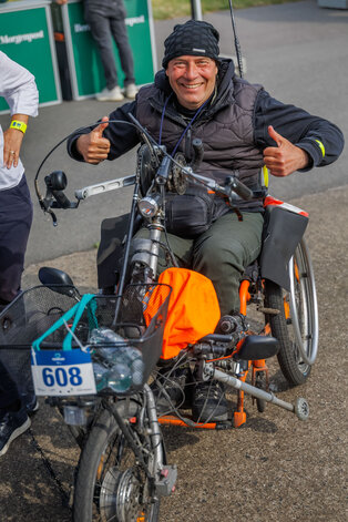 Ein lächelnder Mann sitzt auf einem dreirädrigen Fahrzeug. Er trägt eine schwarze Mütze und eine dunkelgraue Jacke. Mit beiden Daumen zeigt er ein „Daumen hoch“ Zeichen. Im Hintergrund sind verschwommene Personen und eine Veranstaltungseinrichtung zu sehen.