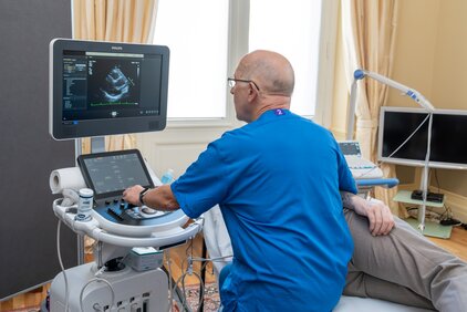 Ein Arzt führt eine Ultraschalluntersuchung durch. Er sitzt vor einem Bildschirm, der Bilder zeigt, während sein Patient auf der Liege liegt. Der Raum hat große Fenster und ist hell und freundlich eingerichtet.