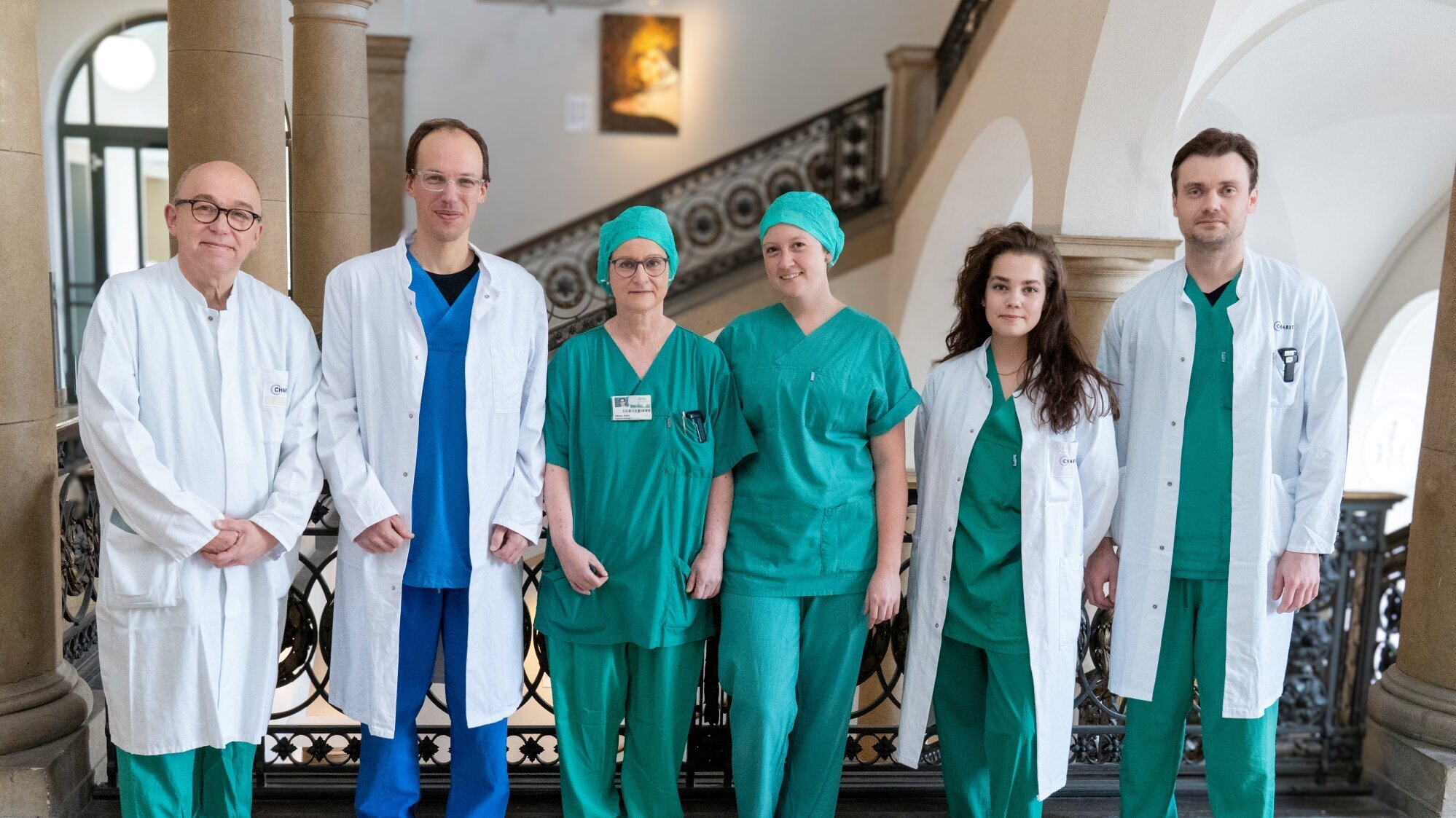 Sechs medizinische Fachkräfte stehen in einem Krankenhausflur. Sie tragen grüne Dienstkleidung und weiße Kittel. Im Hintergrund sind architektonische Elemente des Gebäudes sichtbar. Die Stimmung wirkt professionell und freundlich.