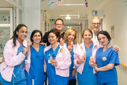 Eine Gruppe von acht medizinischen Fachkräften steht in einem Flur eines Krankenhauses. Sie tragen blaue Arbeitskleidung und lächeln, während sie kleine Schokoladenfiguren in der Hand halten. Der Hintergrund zeigt typisches Krankenhausinterieur mit einem Schild für die Toilette.