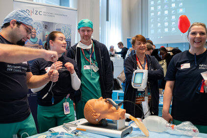 Eine Gruppe von fünf medizinischen Fachkräften steht um ein Simulationsmodell eines menschlichen Kopfes. Sie führen eine Demonstration zur Beatmung durch. Im Hintergrund sind Informationsmaterialien und ein Monitor sichtbar. Die Atmosphäre ist interaktiv und lehrreich.