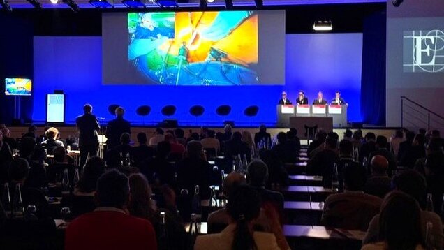 Eine Konferenz oder Veranstaltung im Gange, bei der ein großes Bild auf einer Leinwand gezeigt wird. Im Vordergrund sind mehrere Personen am Rednerpult zu sehen, während das Publikum aufmerksam zuhört und applaudiert. Der Raum ist elegant mit Beleuchtung gestaltet.