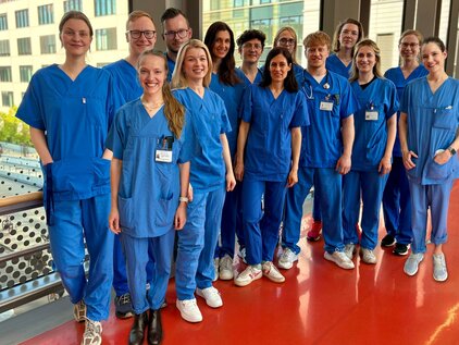 Eine Gruppe von 15 Personen steht nebeneinander in blauen Krankenhausanzügen. Sie lächeln und sind in einem modernen Indoor-Bereich mit großen Fenstern. Einige tragen Namensschilder. Der Boden ist rot, und im Hintergrund sind Gebäude sichtbar.