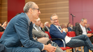 Eine Gruppe von Menschen sitzt auf Stühlen in einem Veranstaltungsraum. Zwei Männer im Vordergrund lachen und unterhalten sich. Der Raum hat eine moderne Ausstattung mit Holzpaneelen und einem roten Hintergrund.