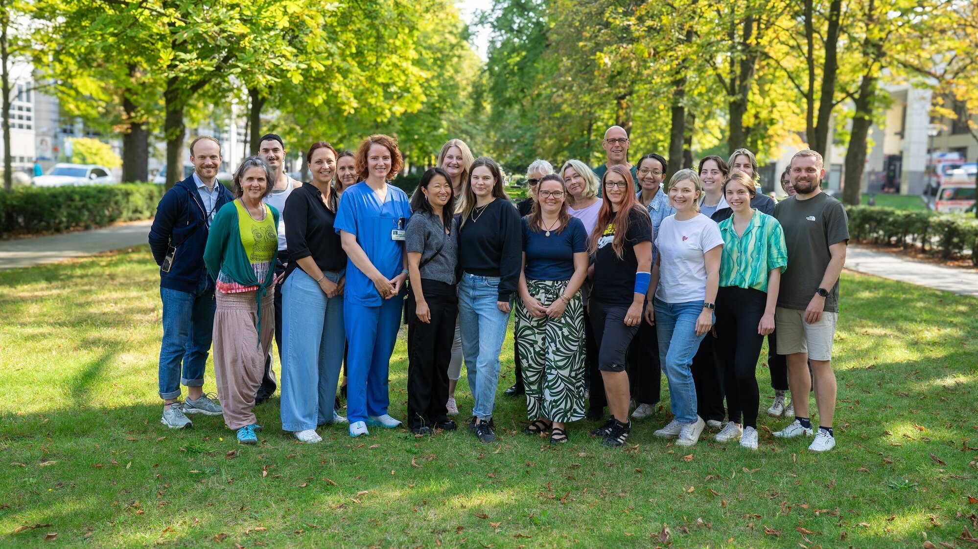 Eine Gruppe von etwa zwanzig Personen steht lächelnd auf einer Wiese, umgeben von Bäumen. Sie tragen verschiedene, bunte Kleidung und scheinen freundlich und entspannt. Der Hintergrund zeigt gepflegte Grünflächen und einige Gebäude im Hintergrund.