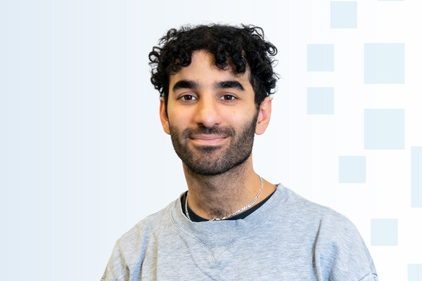 Ein junger Mann mit lockigem, schwarzem Haar lächelt in die Kamera. Er trägt ein graues Sweatshirt und hat einen schlichten Schmuckanhänger um den Hals. Der Hintergrund ist hell und enthält einige geometrische Formen.