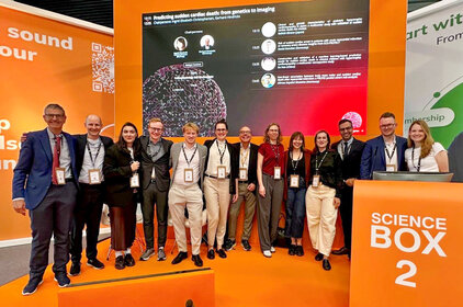 Eine Gruppe von zwölf Personen steht vor einer orangefarbenen Wand mit Informationen zur Präsentation "Vorhersage von Herzkrankheiten durch genetische Analyse". Alle Teilnehmer tragen Namensschilder und lächeln, während sie auf das Thema der wissenschaftlichen Veranstaltung fokussiert sind.