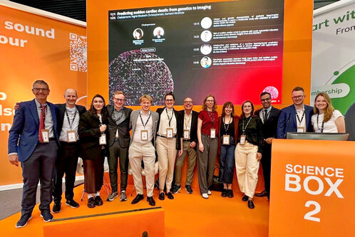 Eine Gruppe von zwölf Personen steht vor einer orangefarbenen Wand mit Informationen zur Präsentation "Vorhersage von Herzkrankheiten durch genetische Analyse". Alle Teilnehmer tragen Namensschilder und lächeln, während sie auf das Thema der wissenschaftlichen Veranstaltung fokussiert sind.
