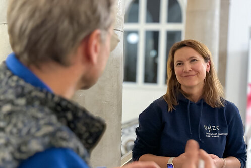 Zwei Personen unterhalten sich in einem Raum. Eine Frau lächelt und trägt einen blauen Hoodie, während der Blick auf einen Mann gerichtet ist, der mit seiner Hand gestikuliert. Im Hintergrund sind unscharfe Elemente und Fenster zu sehen.