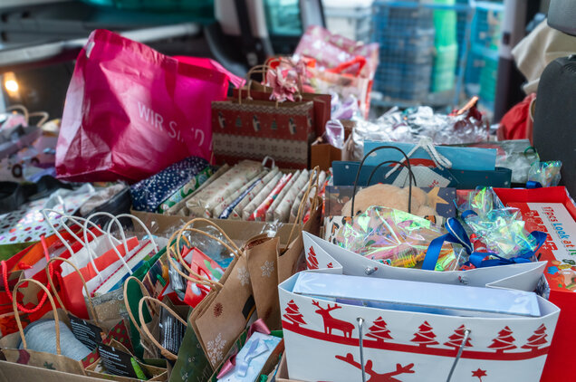 Ein Auto ist voller bunt verpackter Geschenke in verschiedenen Taschen und Kästen. Die Geschenke sind weihnachtlich dekoriert, mit bunten Schleifen und unterschiedlichen Mustern. Im Hintergrund sind weitere Taschen und Geschenke sichtbar.