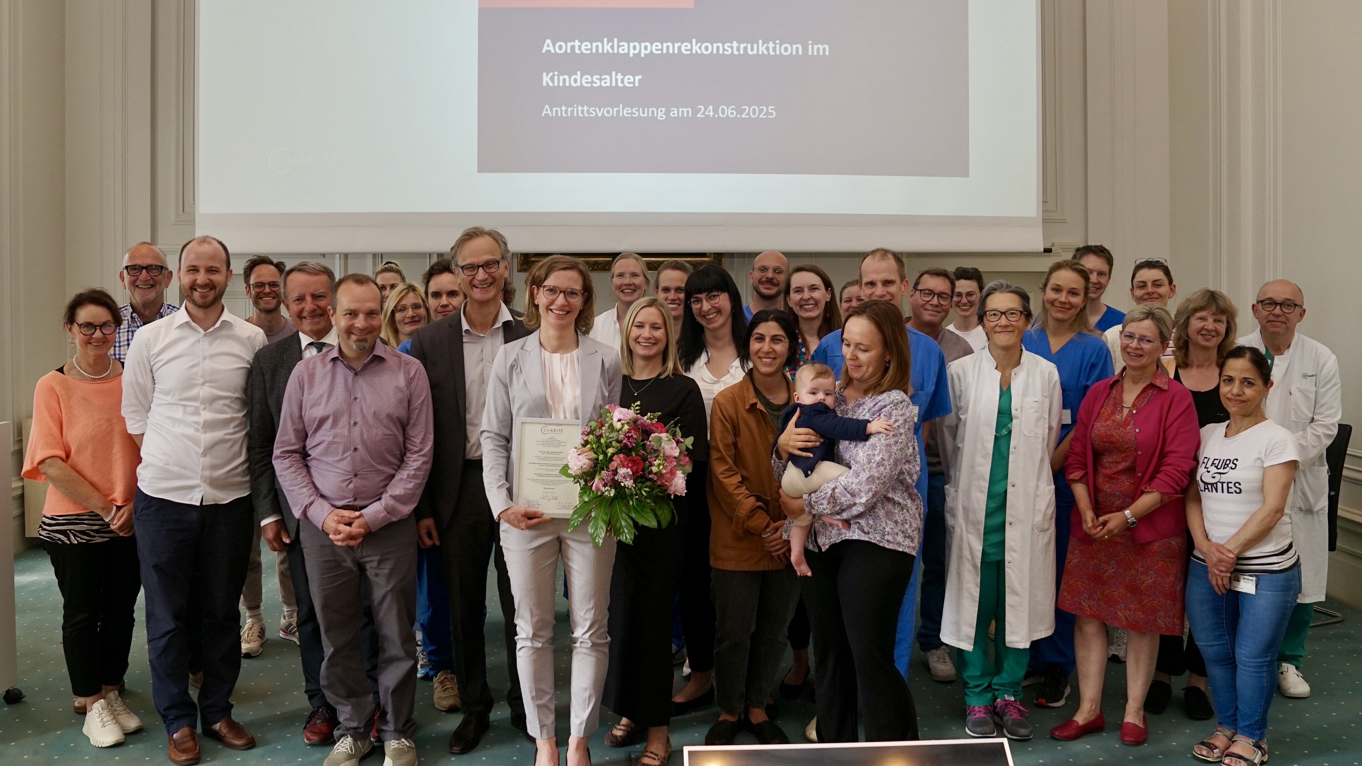 Eine Gruppe von über 30 Personen posiert zu einer Feier. In der Mitte steht eine Frau mit einem Zertifikat und einem Blumenstrauß. Sie trägt einen hellen Anzug und lächelt. Im Hintergrund ist eine Präsentation zum Thema „Aortenklappenrekonstruktion im Kindesalter“ sichtbar.