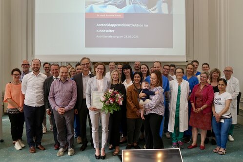 Eine Gruppe von über 30 Personen posiert zu einer Feier. In der Mitte steht eine Frau mit einem Zertifikat und einem Blumenstrauß. Sie trägt einen hellen Anzug und lächelt. Im Hintergrund ist eine Präsentation zum Thema „Aortenklappenrekonstruktion im Kindesalter“ sichtbar.