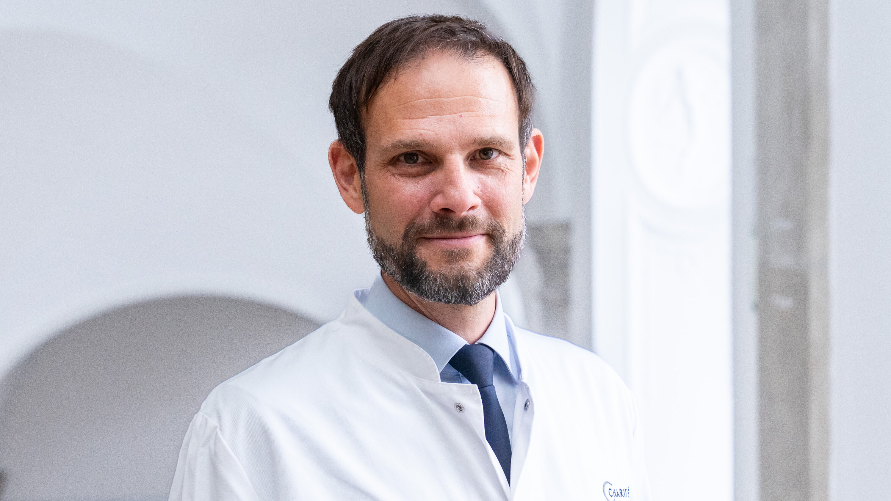 Ein männlicher Arzt steht lächelnd vor einem hellen, minimalistischen Hintergrund. Er trägt einen weißen Kittel über einem Hemd und einer Krawatte und hat einen gepflegten Bart. Sein Ausdruck wirkt freundlich und kompetent.