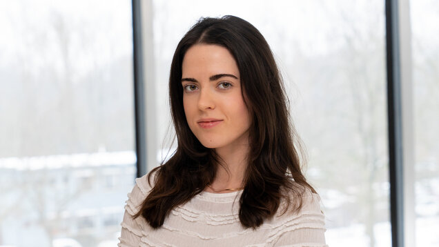 Eine junge Frau mit langen, dunklen Haaren und einem selbstbewussten Ausdruck sitzt vor einem großen Fenster. Die Umgebung ist hell und winterlich. Sie trägt ein helles, strukturiertes Oberteil.