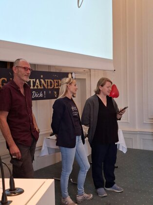 Drei Personen stehen auf der Bühne in einem eleganten Raum. Die Frau in der Mitte hält ein Handy in der Hand, während die anderen beiden entspannt in ihre Richtung schauen. Im Hintergrund ist ein Banner mit dem Text "Herzlich Willkommen!" sichtbar.