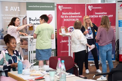 Auf der Veranstaltung interagieren mehrere Frauen an Ständen, die Informationen über Gesundheitsangebote bereitstellen. Ein Banner informiert über die „Fontanherzen-App“ und einen nationalen Service für Herzfehler. Die Atmosphäre ist lebhaft, mit Gesprächen und Informationsaustausch im Vordergrund.
