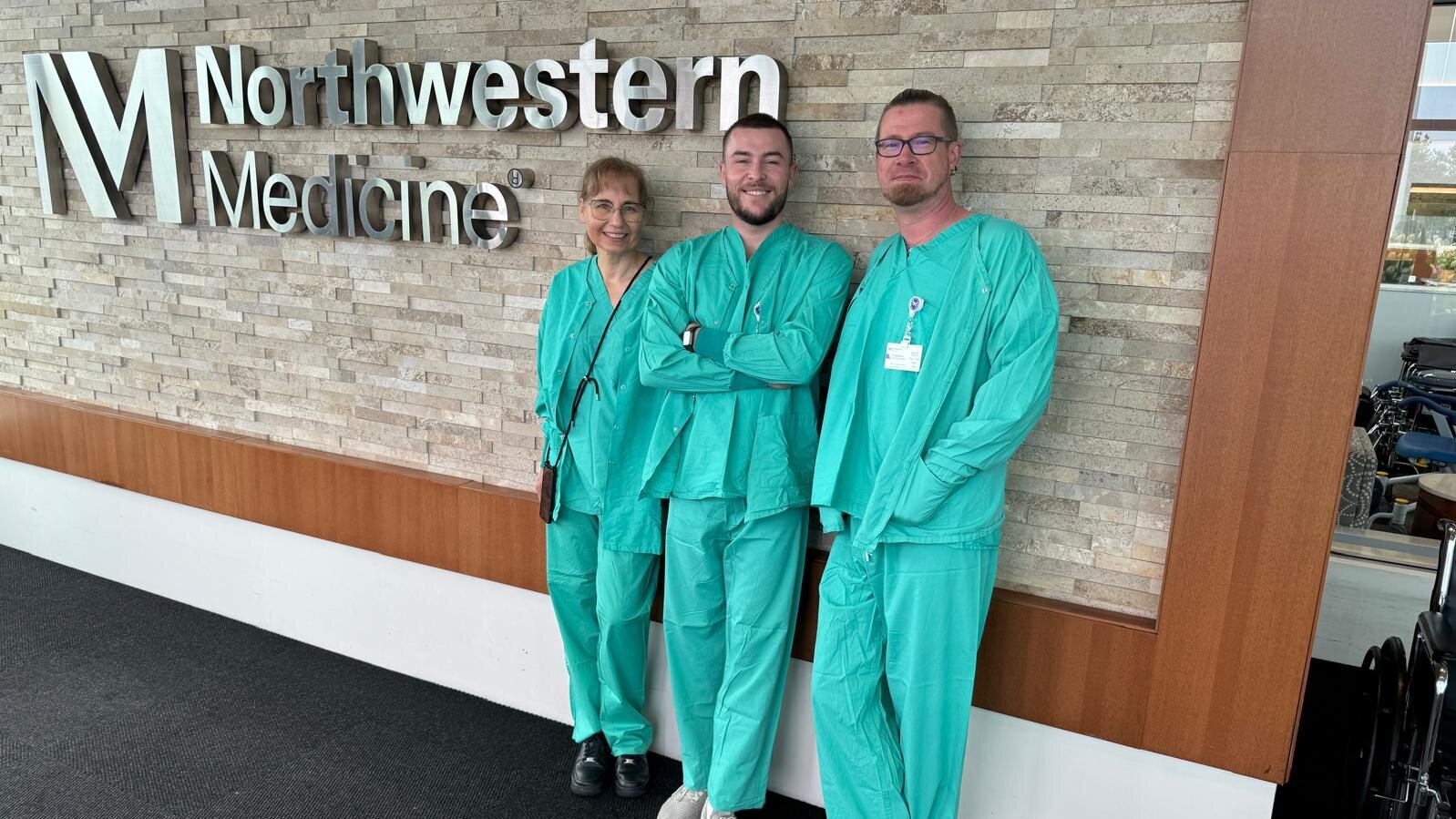Drei Personen in grünen OP-Kitteln stehen lächelnd vor einer Wand mit dem Schriftzug "Northwestern Medicine". Die Umgebung hat eine moderne, freundliche Atmosphäre.