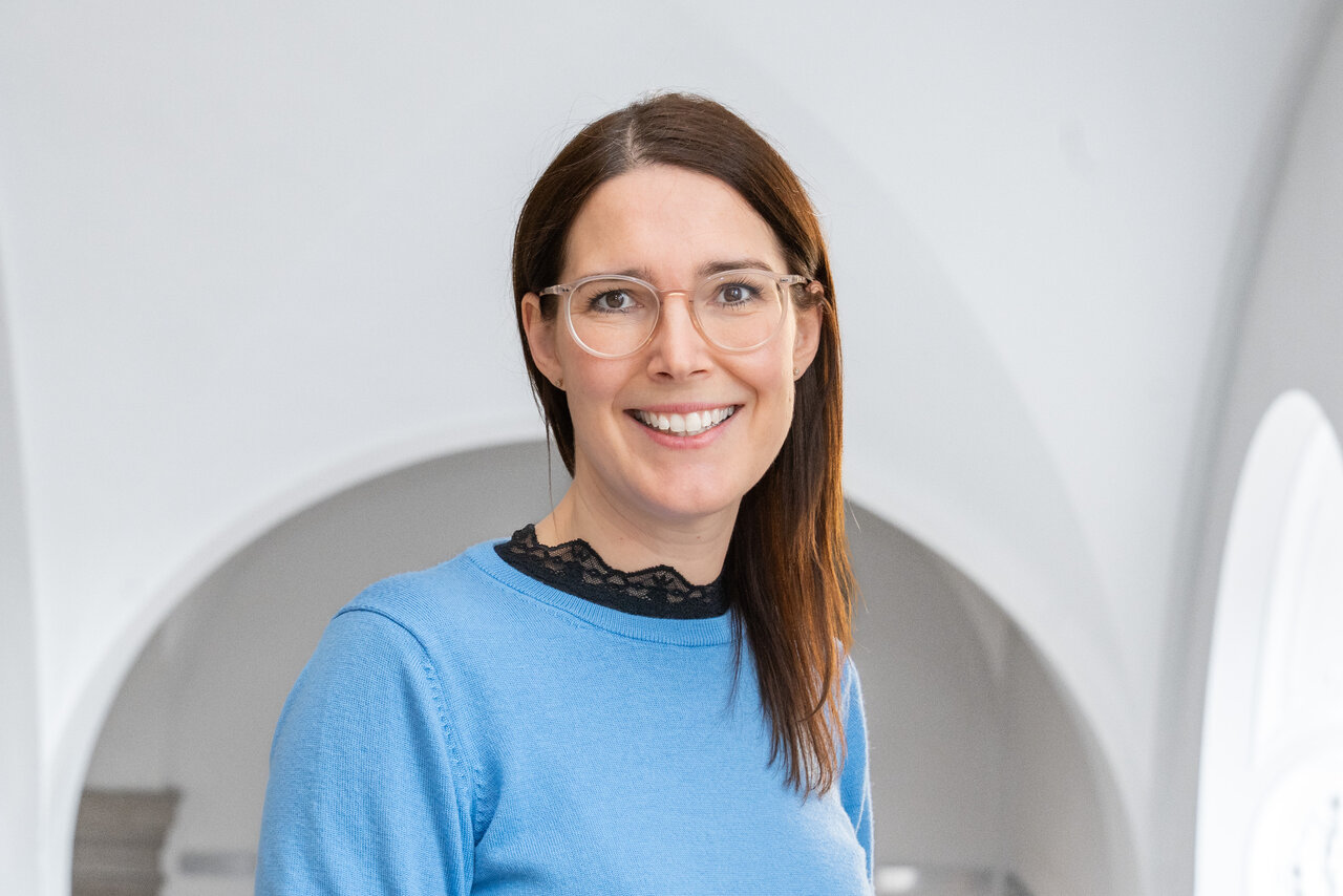 Ein Porträt einer lächelnden Frau mit langen, braunen Haaren, die eine blaue Bluse und eine schwarze Spitze trägt. Sie trägt eine Brille und steht vor einem hellen, neutralen Hintergrund. Die Frau wirkt freundlich und zugänglich.