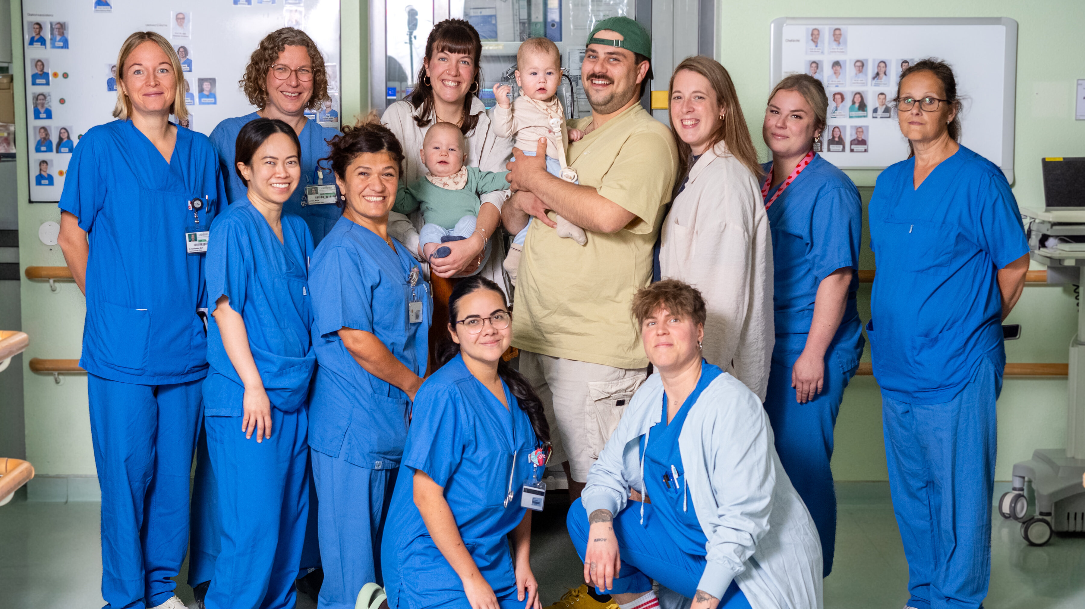 Eine Gruppe von elf medizinischen Fachkräften in blauen Uniformen steht in einem Krankenhauszimmer. In der Mitte hält eine Person ein Baby, während ein weiteres Baby in den Armen einer anderen Person sitzt. Die Atmosphäre wirkt freundlich und unterstützend.