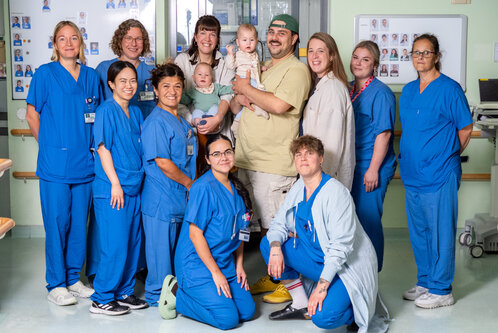 Eine Gruppe von elf medizinischen Fachkräften in blauen Uniformen steht in einem Krankenhauszimmer. In der Mitte hält eine Person ein Baby, während ein weiteres Baby in den Armen einer anderen Person sitzt. Die Atmosphäre wirkt freundlich und unterstützend.