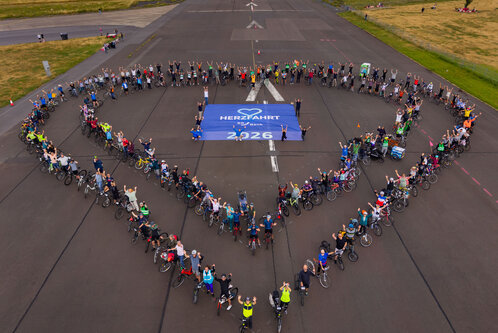 Eine große Gruppe von Menschen steht auf einer Landebahn und bildet mit Fahrrädern ein Herz. In der Mitte des Herzens ist ein Banner mit der Aufschrift „HERZLICH WILLKOMMEN 2026“. Der Himmel ist bewölkt und die Umgebung zeigt grünes Gras.